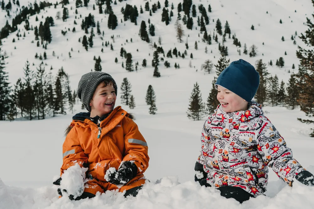 Zwei Kinder sitzen lachend im Schnee vor einem verschneiten, mit Tannen bewachsenen Hang