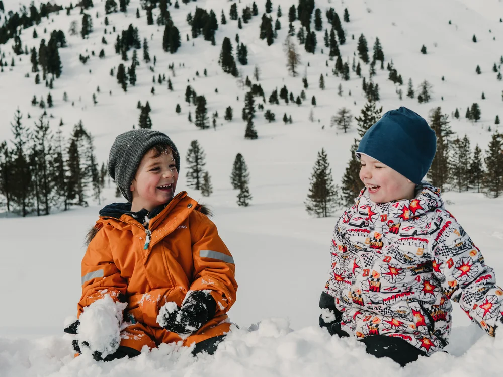 GRETI in Bad Hofgastein: dein alpines Boutique Hotel Zwei Kinder sitzen lachend im Schnee vor einem verschneiten, mit Tannen bewachsenen Hang