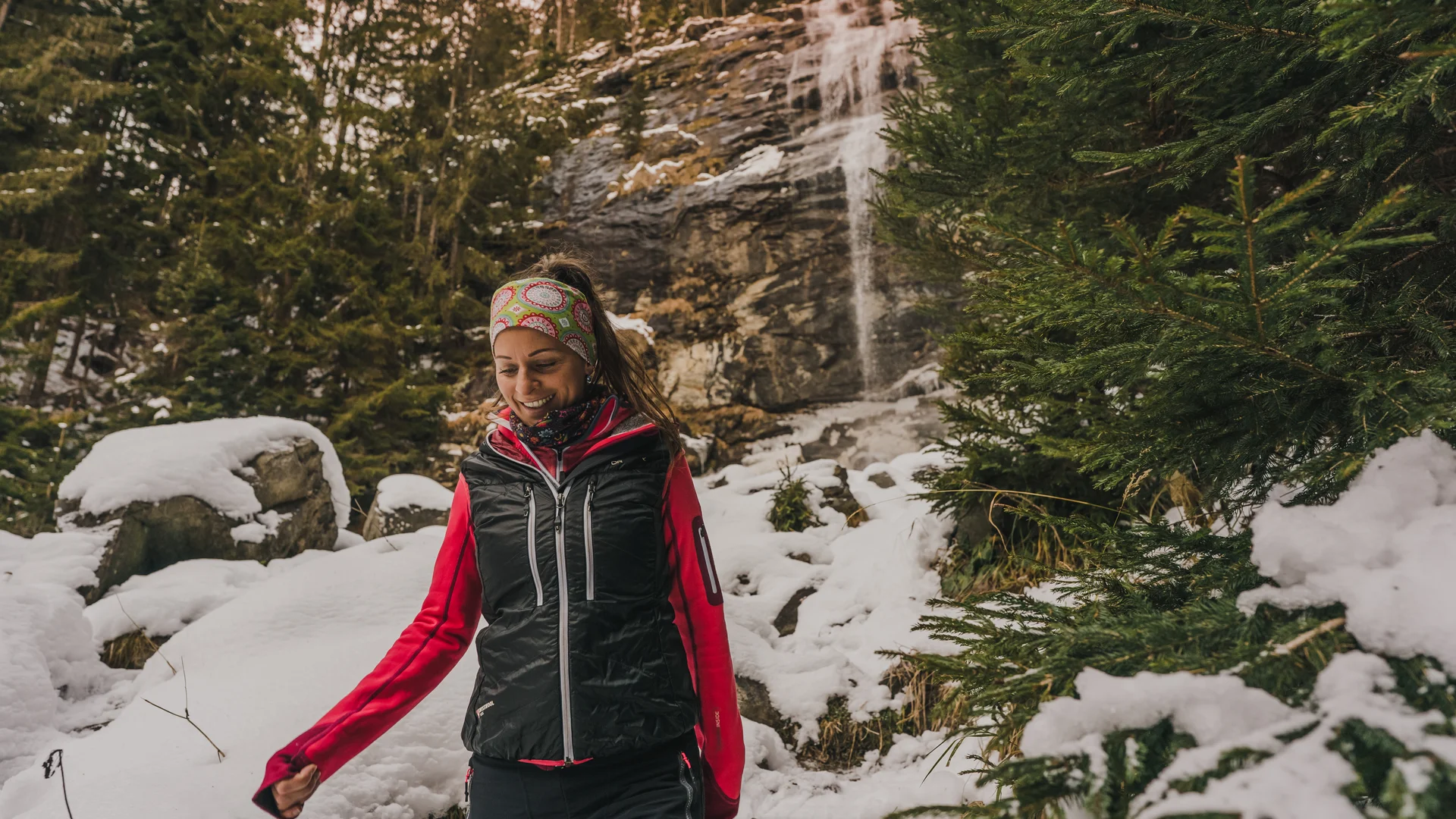 Lächelnde Frau in Winterkleidung an einem Wasserfall im verschneiten Wald